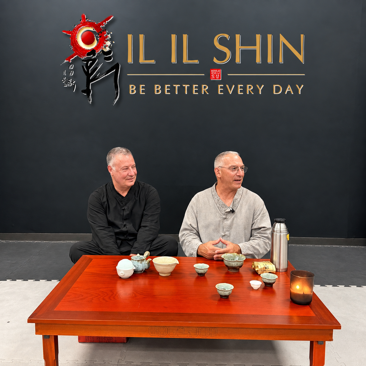 Two people sharing tea beneath the Il Il Shin — Be Better Every Day wall emblem.